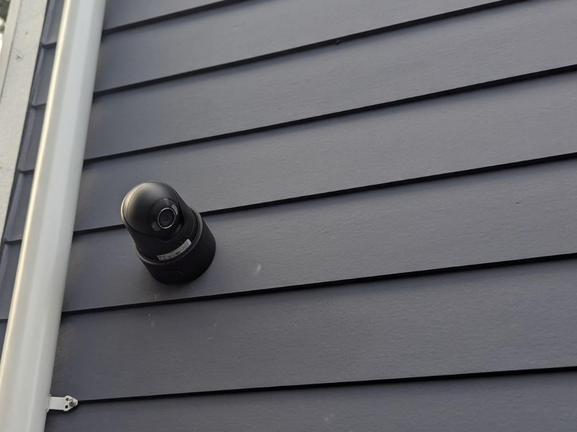 A black Ajax turret camera mounted on dark gray lap siding beside a white downspout