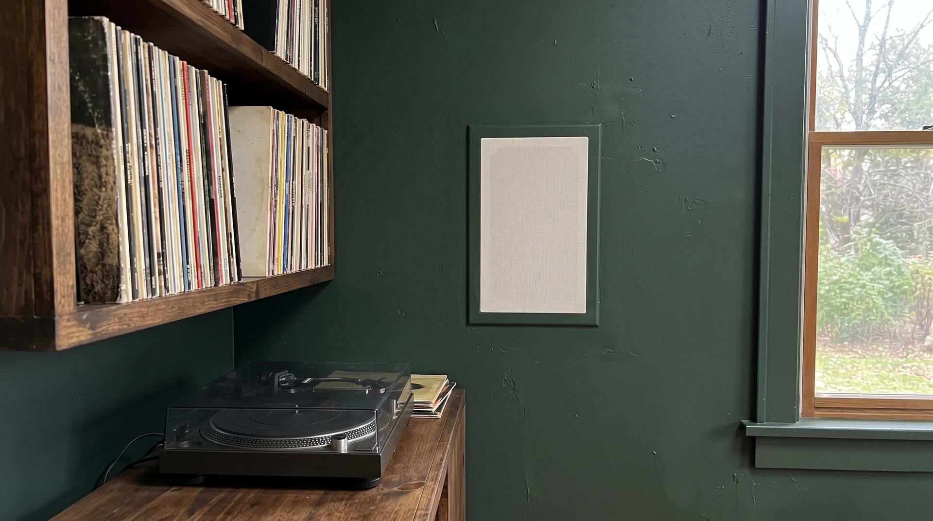 A flush in-wall speaker with a plain grille in a dark green wall beside a record shelf and turntable