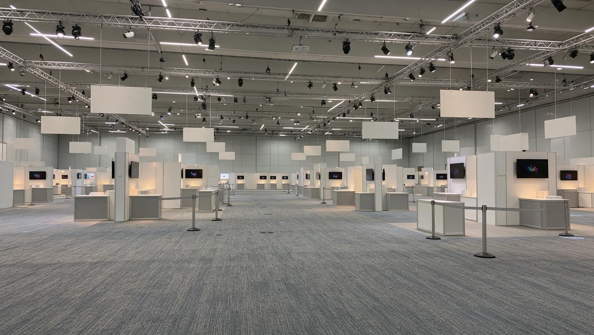 An empty trade show hall with exhibit booths, wall-mounted TVs and hanging signs under steel trusses