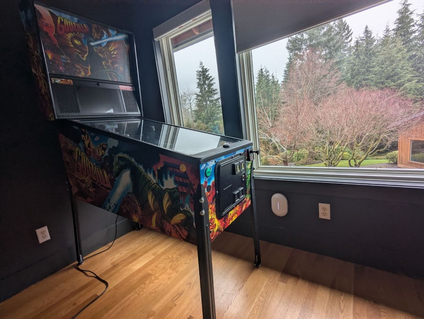 A white Alta Labs Summit-A6 Wi-Fi 6 access point mounted low on a dark wall under a window in a North Bend game room, beside a pinball machine