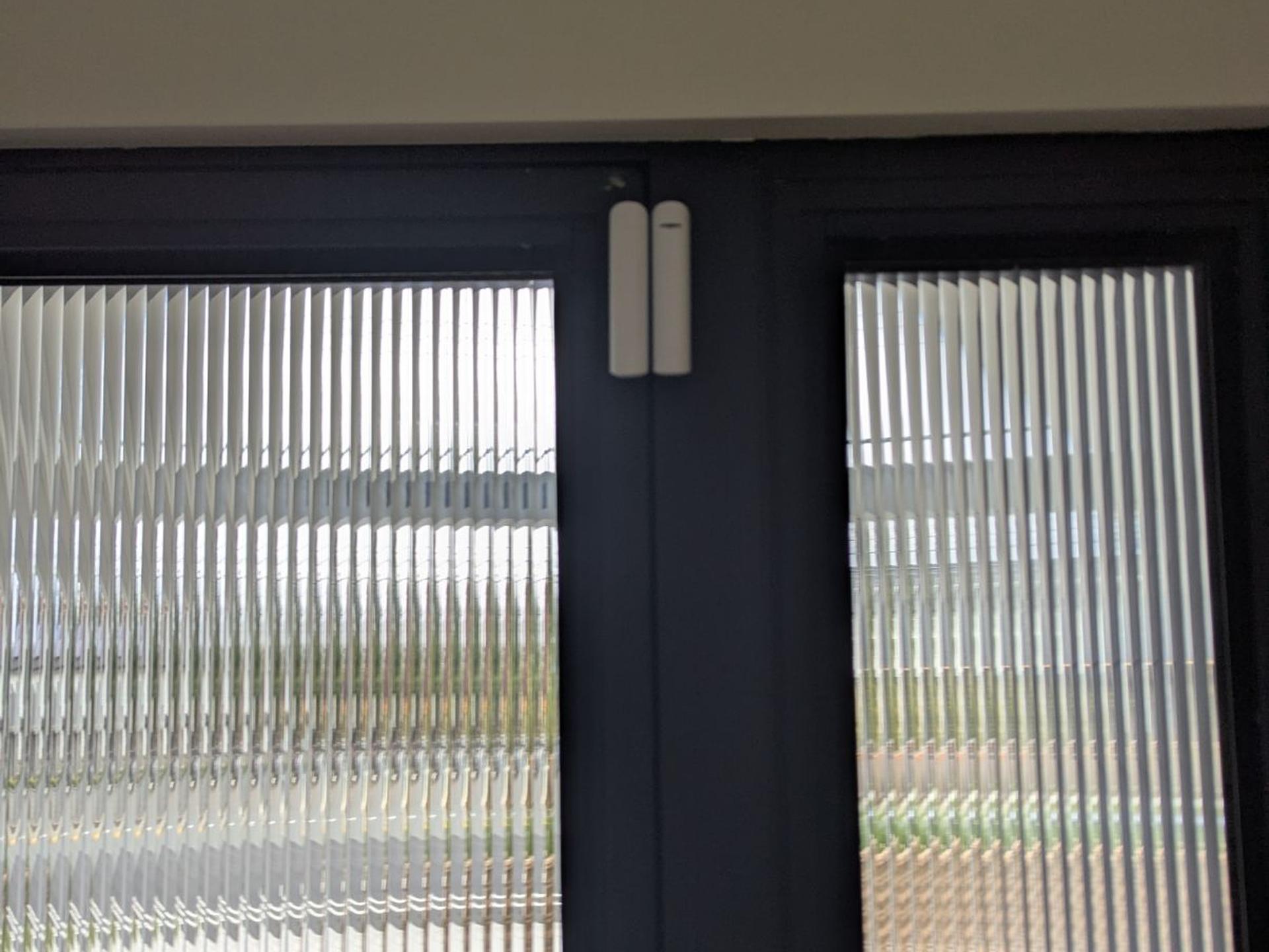 A white Ajax DoorProtect sensor at the top of a black-framed door with reeded glass panels