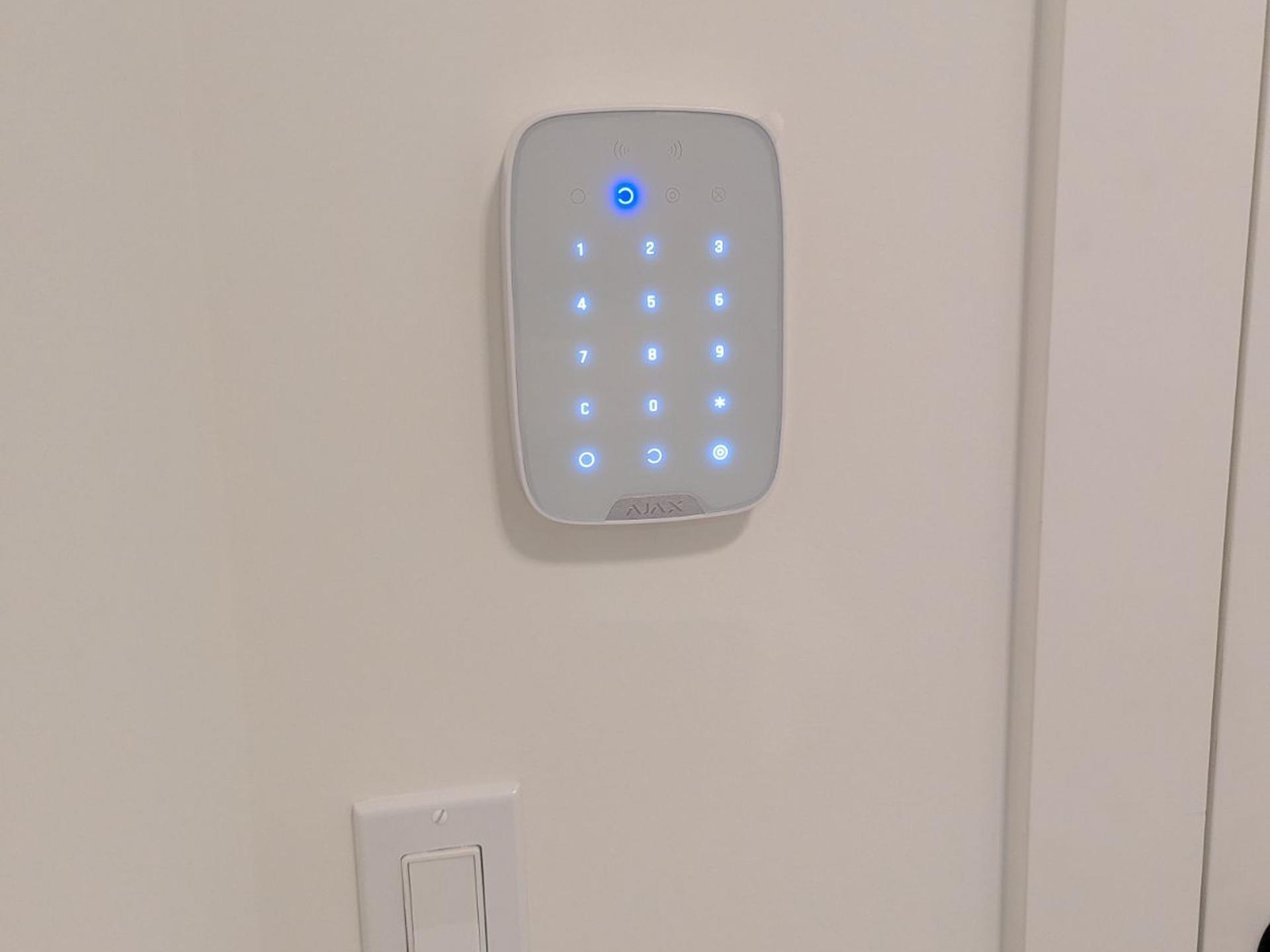 A white Ajax KeyPad Plus lit blue on a cream wall above a light switch