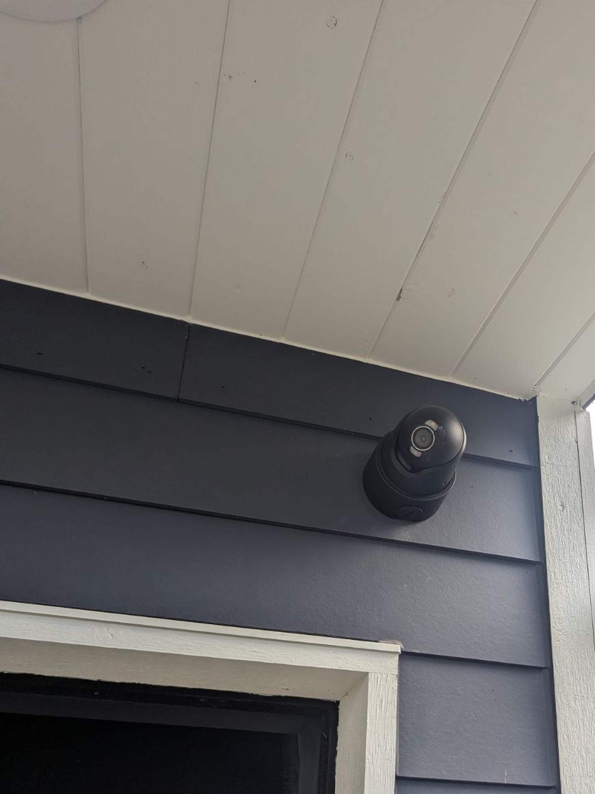 A black Ajax turret camera under a white porch ceiling, above a garage door, on dark gray siding