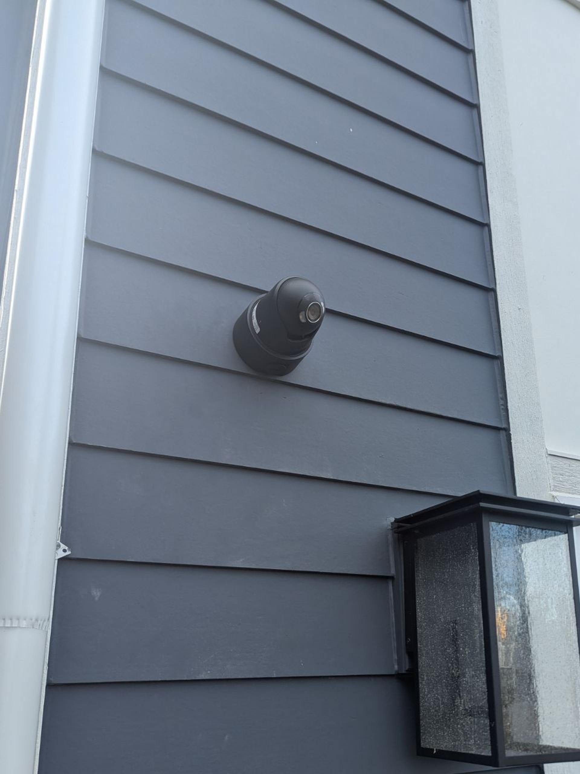 A black Ajax turret camera on dark gray siding above a black lantern-style porch light