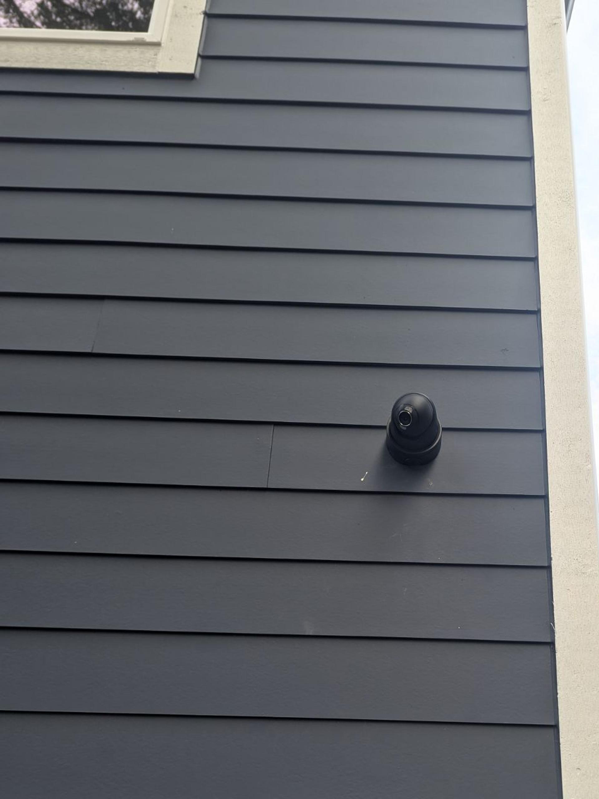 A black Ajax turret camera high on a dark gray side wall below an upstairs window