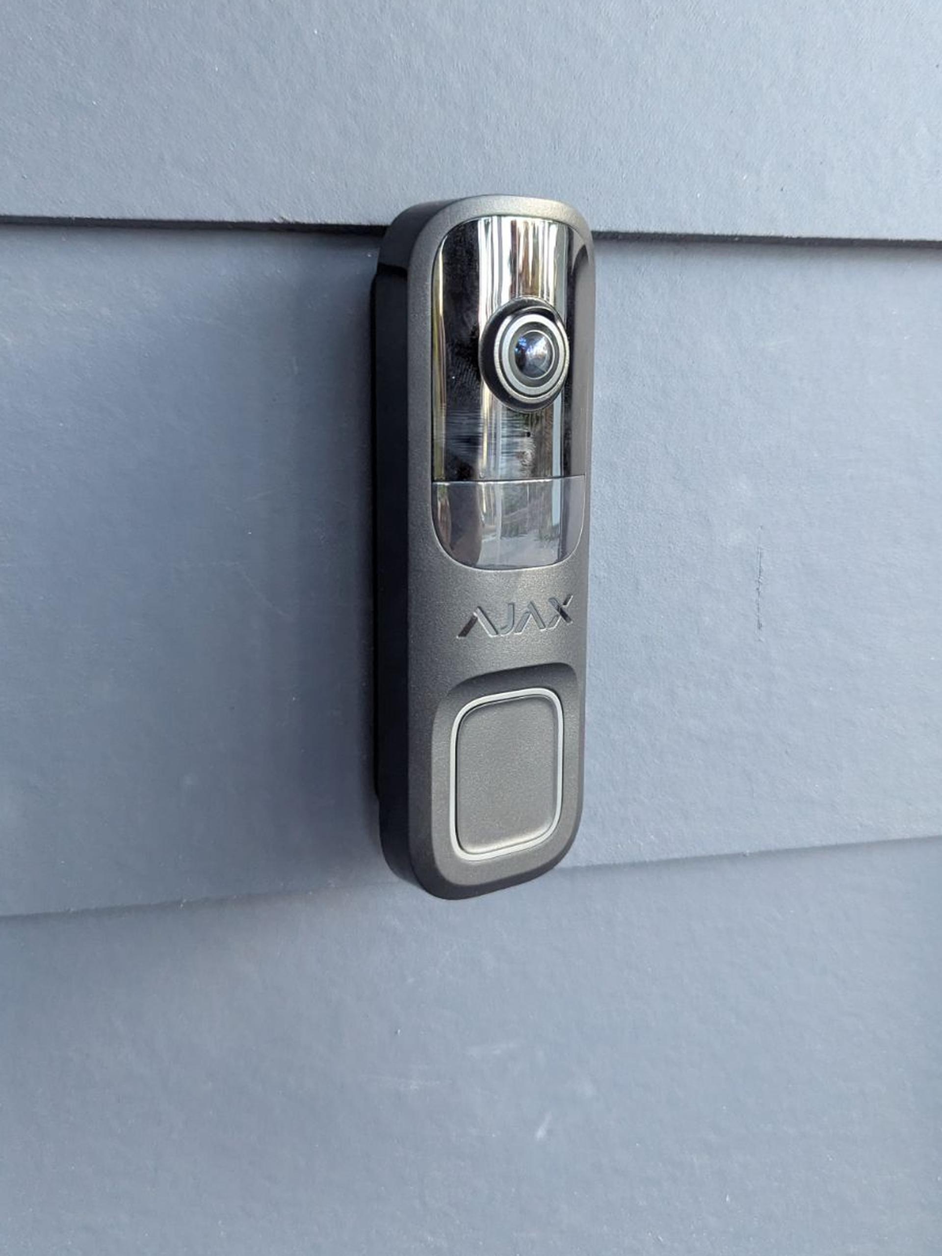 A black Ajax video doorbell mounted on gray siding